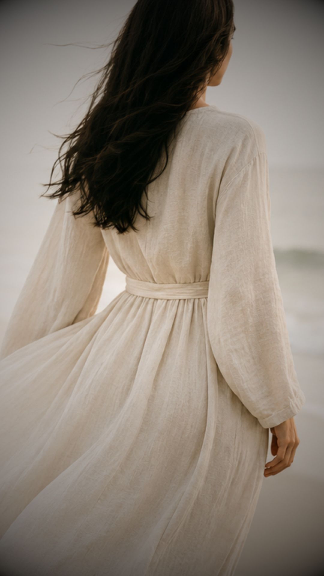 A woman in a flowing cream dress walks along the shore, hair caught in the wind.